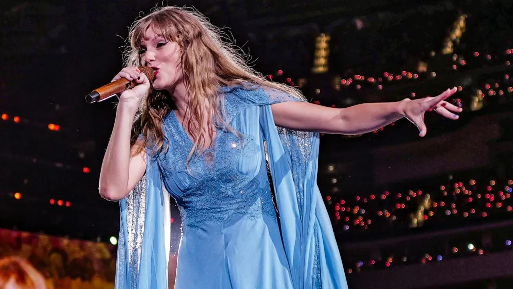 Taylor Swift z wyciągniętą ręką i trzymającą mikrofon podczas koncertu