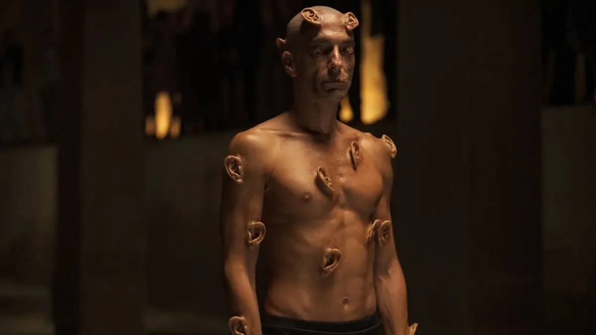 A naked man stands with ears growing out of his body in