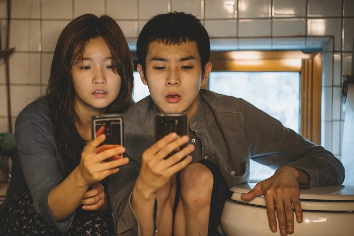 Parasite characters view their devices in front of a toilet in their underground apartment.