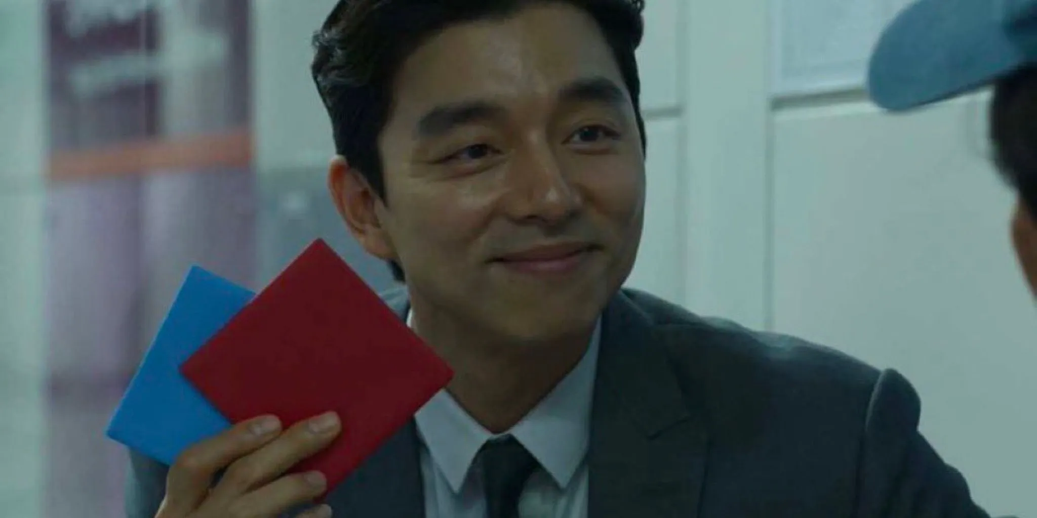 Gong Yoo como O Vendedor segurando uma peça vermelha Ddakji em Squid Game (2021)