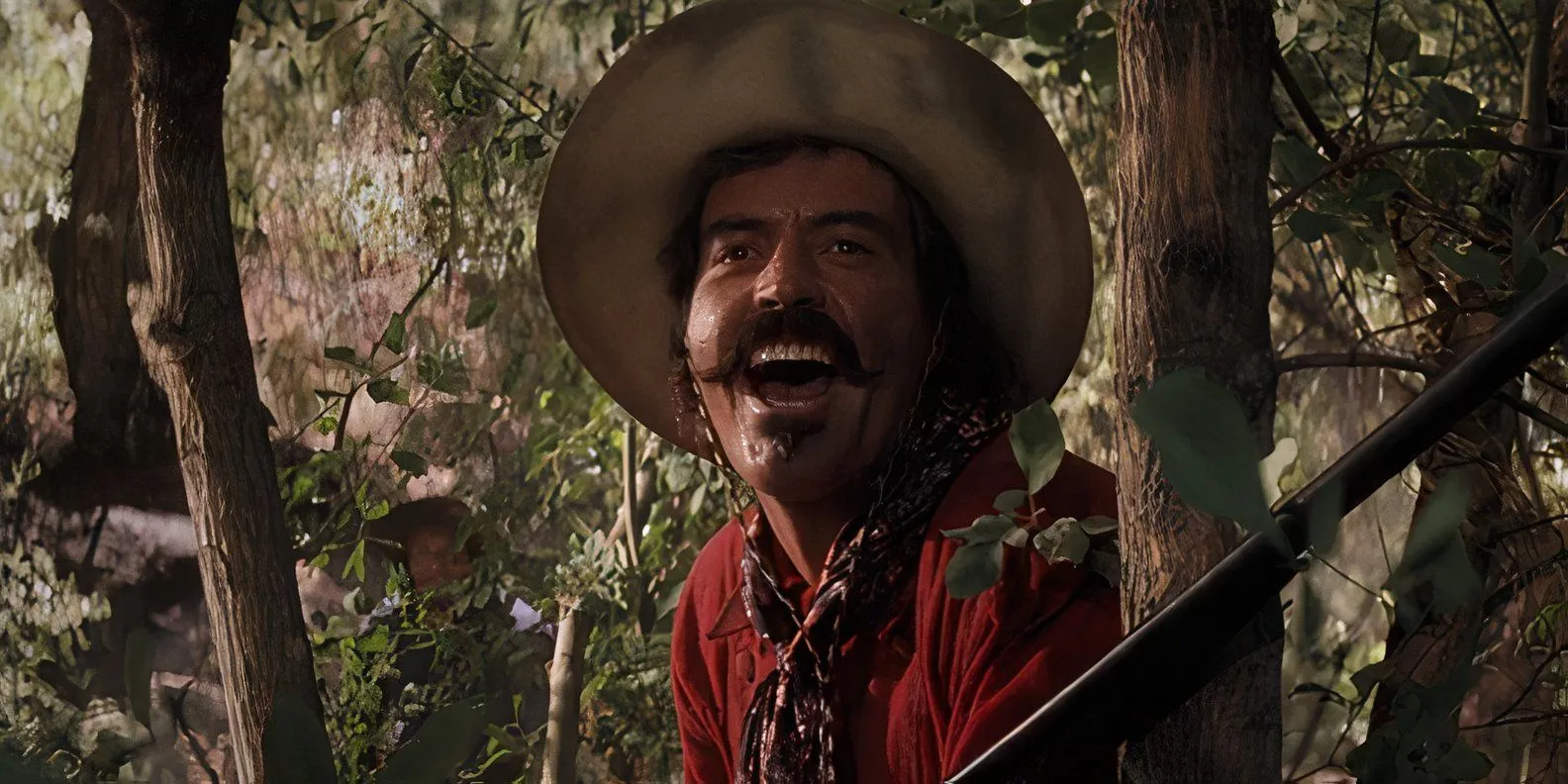 Curly Bill Brocius (Powers Boothe) glousse en se cachant derrière un arbre et en tenant un fusil de chasse à Tombstone