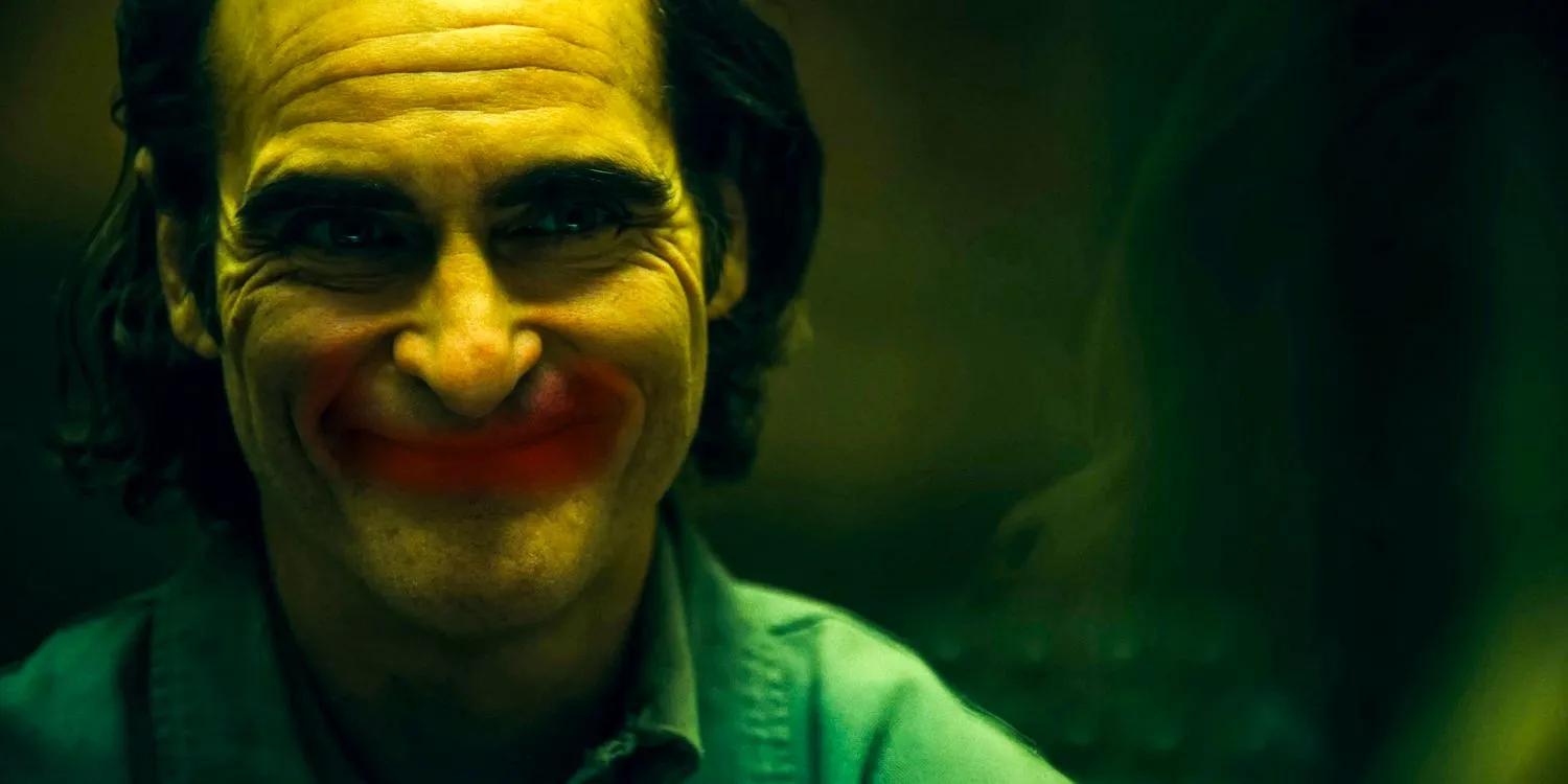 Arthur Fleck (Joaquin Phoenix) con un sorriso forzato si allinea perfettamente con un sorriso disegnato con rossetto rosso su un bicchiere in Joker: Folie à Deux