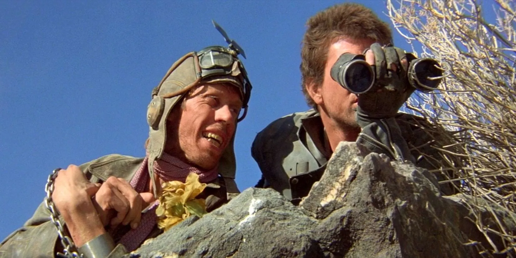 A still image from Mad Max 2: The Road Warrior. Max, played by Mel Gibson, looks through binoculars as the Gyro Captain watches.