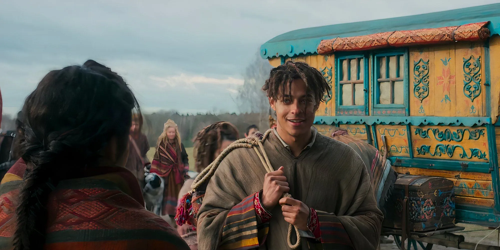 Aram stands in front of a colorful Tuatha'an wagon in The Wheel of Time Aram stands in front of a colorful Tuatha'an wagon in The Wheel of Time