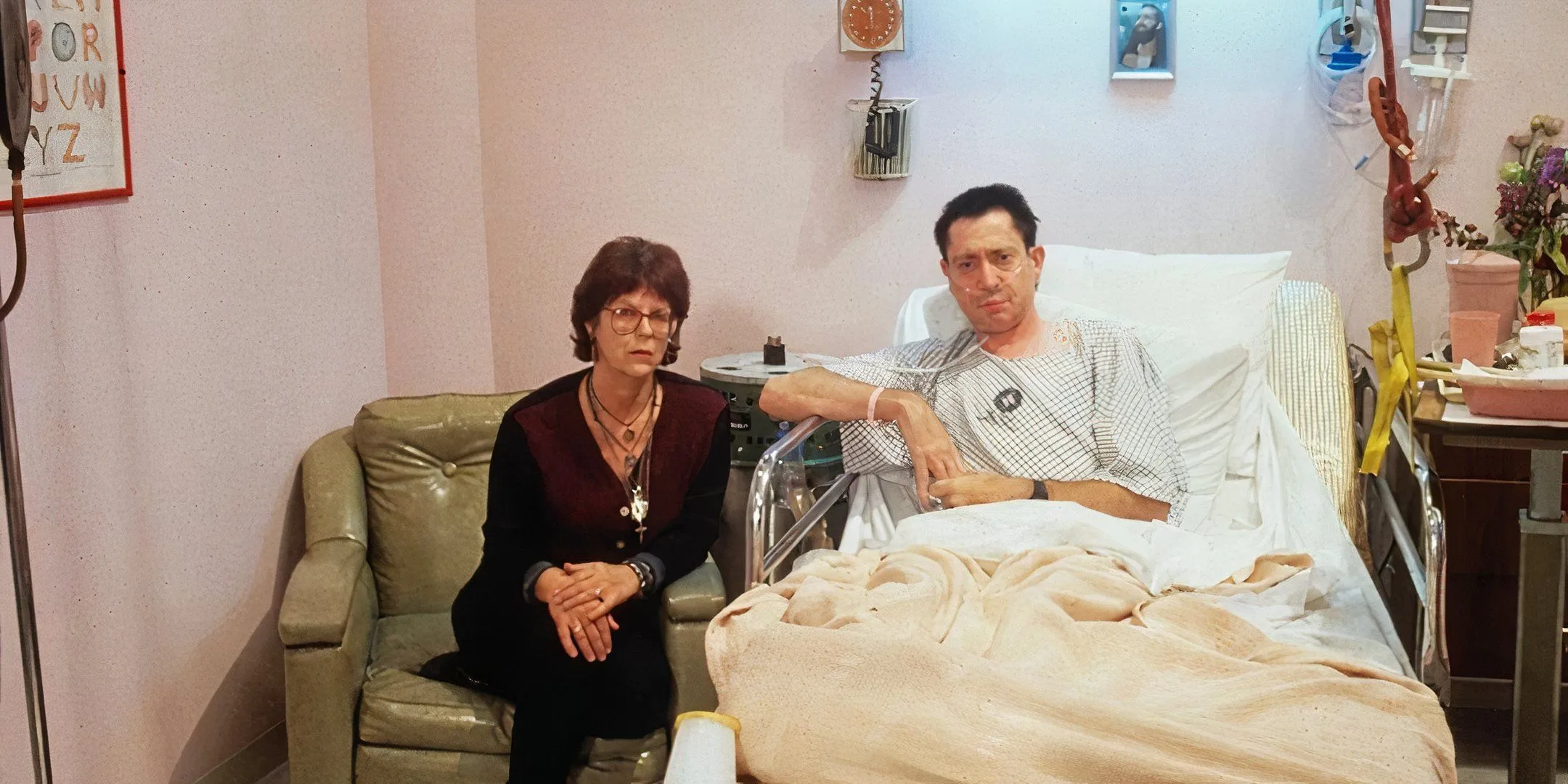 A woman looks at the camera with a serious expression as she sits next to a man in a hospital bed. A woman looks at the camera with a serious expression as she sits next to a man in a hospital bed.